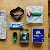 Power Cut Basics Kit laid out on a kitchen table: headlamp, water purification tablets, collapsible jerry can, waterproof matches, foil blanket and Seven Oceans ration block