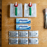 Water at Home Kit on a kitchen table: two folded 10 litre jerry cans, two boxes of Aquatabs and six PainsWessex emergency drinking water sachets